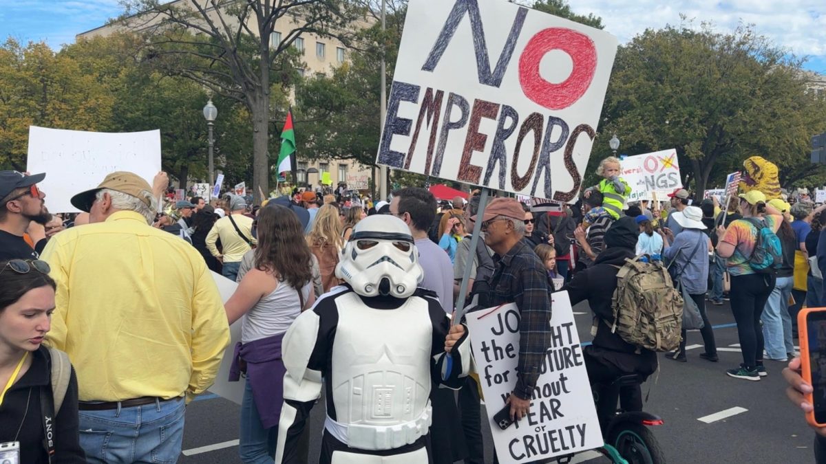 A person costumed as a stormtrooper from Star Wars carries a sign reading "No Emperors."
