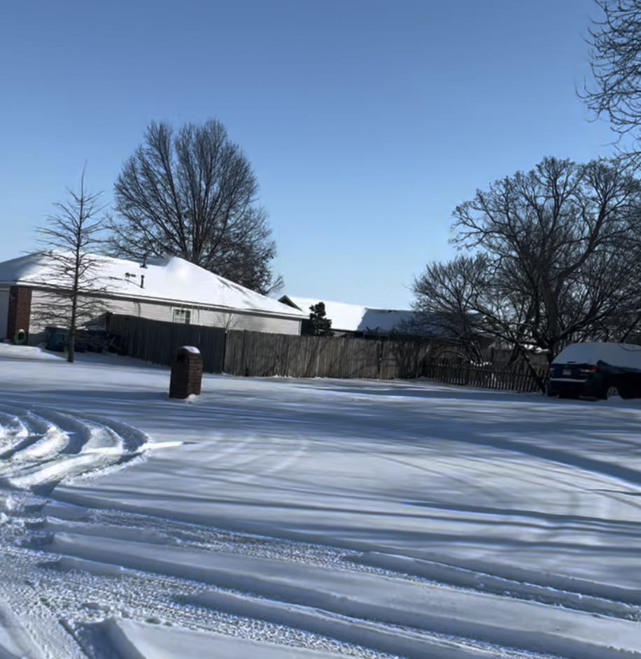 The image shows a snow-covered neighborhood with a few tracks in a roadway. A house is in the background.