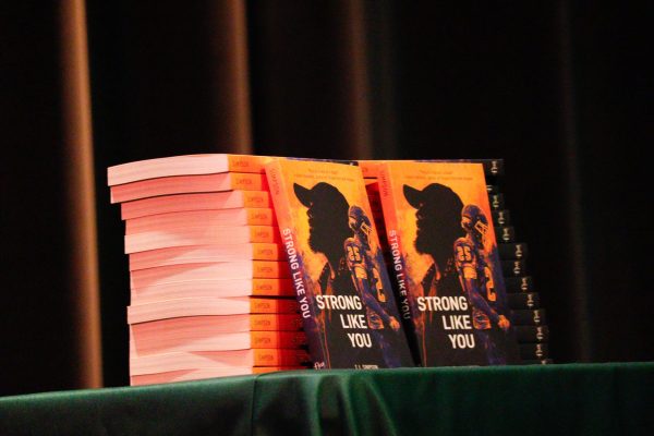 Copies of a book are stacked on a table. A couple of the books are standing up straight in front of the book, which is titled "Strong Like You."