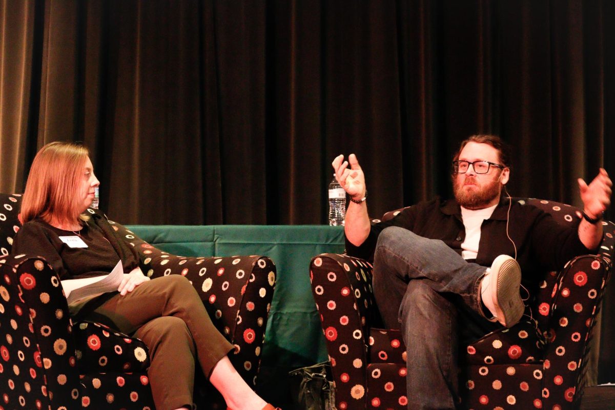 Two people are seated on comfortable chairs in front of an audience. The person on the right is moving his arms.