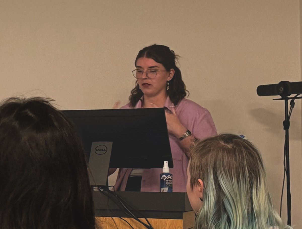 A woman stands behind a computer monitor as she speaks to an audience.
