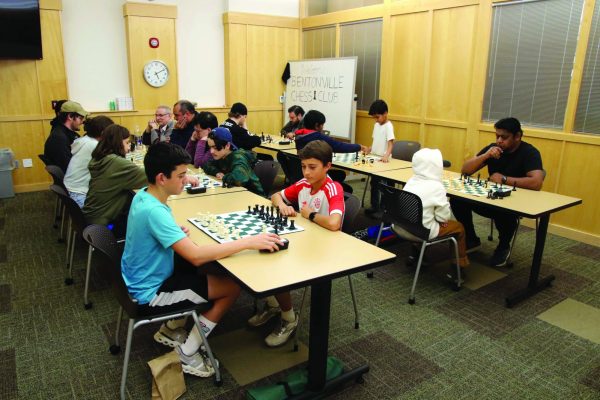 A group of people are seated at tables with chess boards between them.