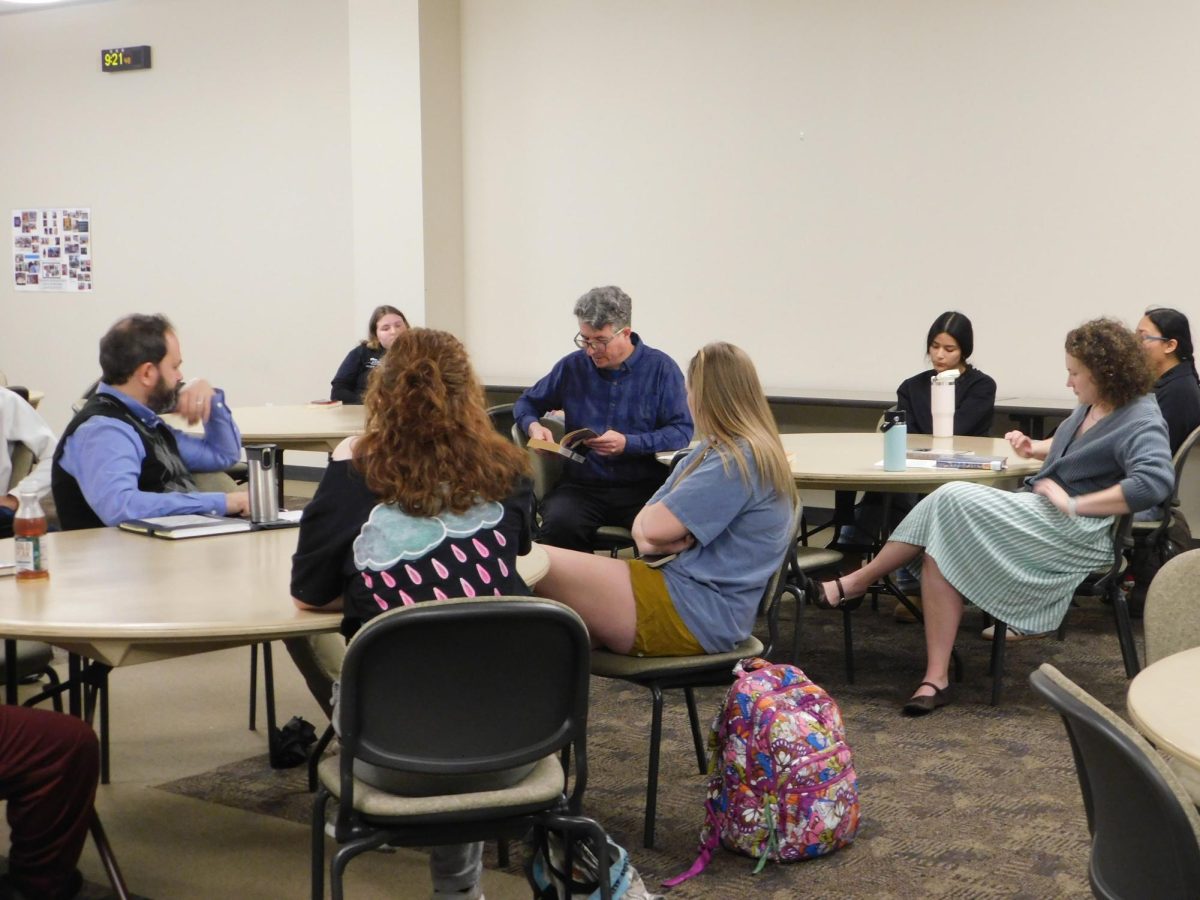 A group of people are seated at round tables with their attention focused on a man holding a book and reading from it.