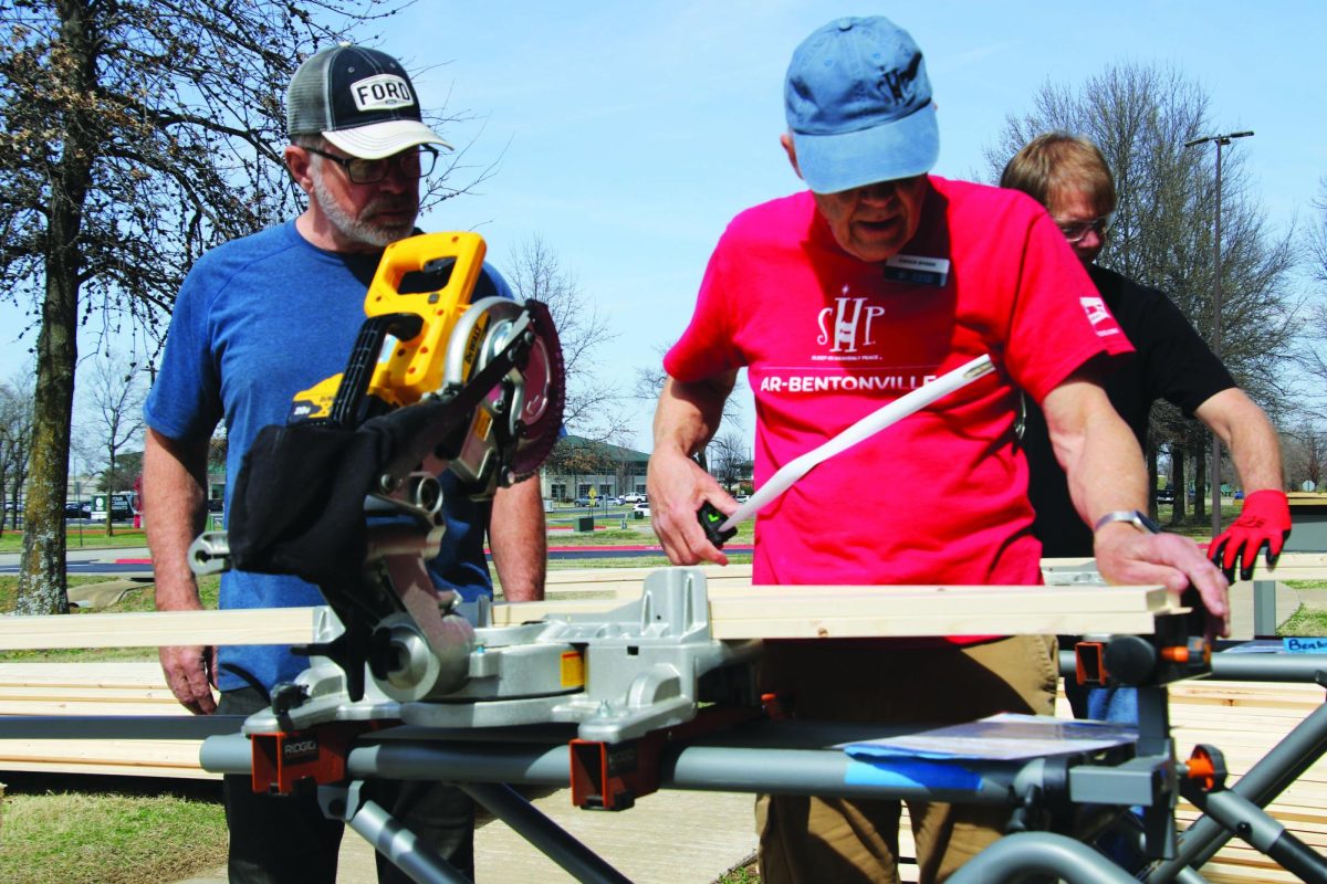 Two men stand behind a piece of lumber, and one of the men is holding a measuring device.