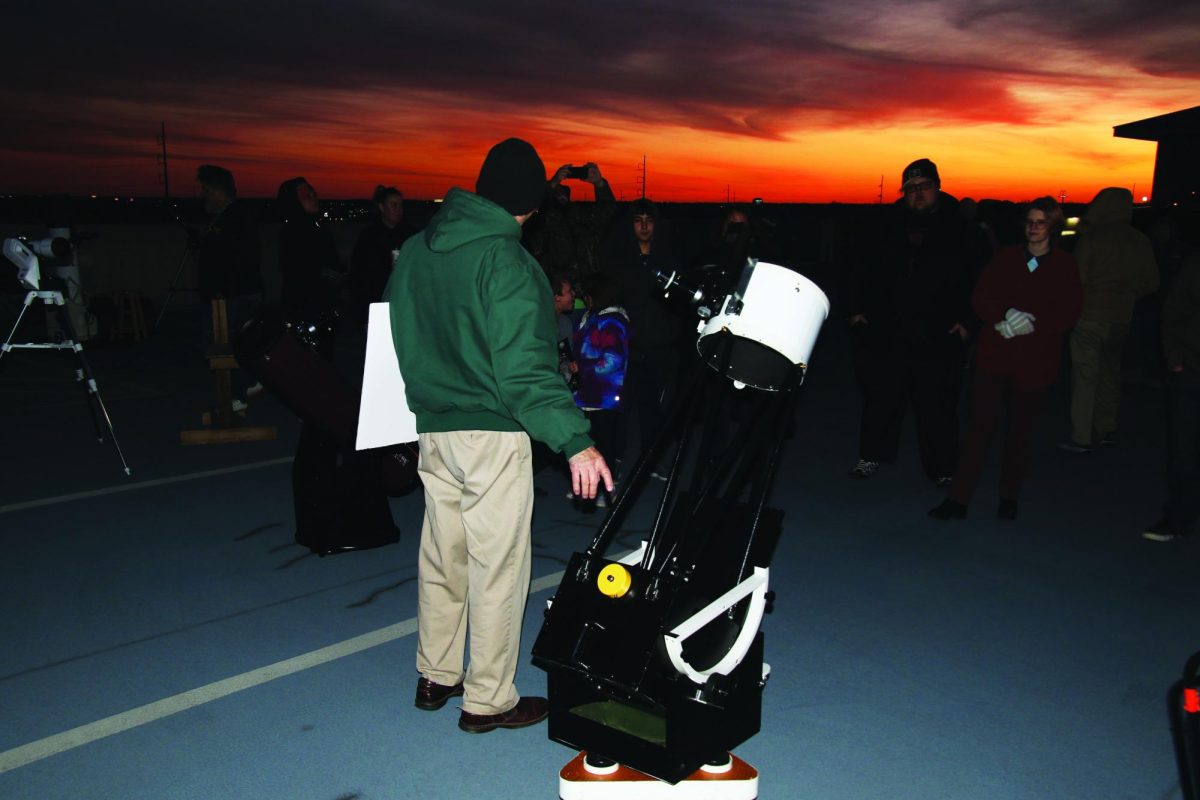 A colorful sunset is visible in the background as stargazers gather around a powerful telescope.