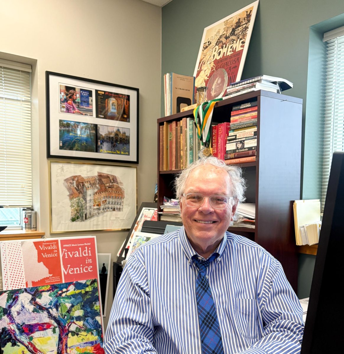 A man wearing a striped shirt and tie sits in an office. Visible behind him is a poster for "Vivaldi in Venice"