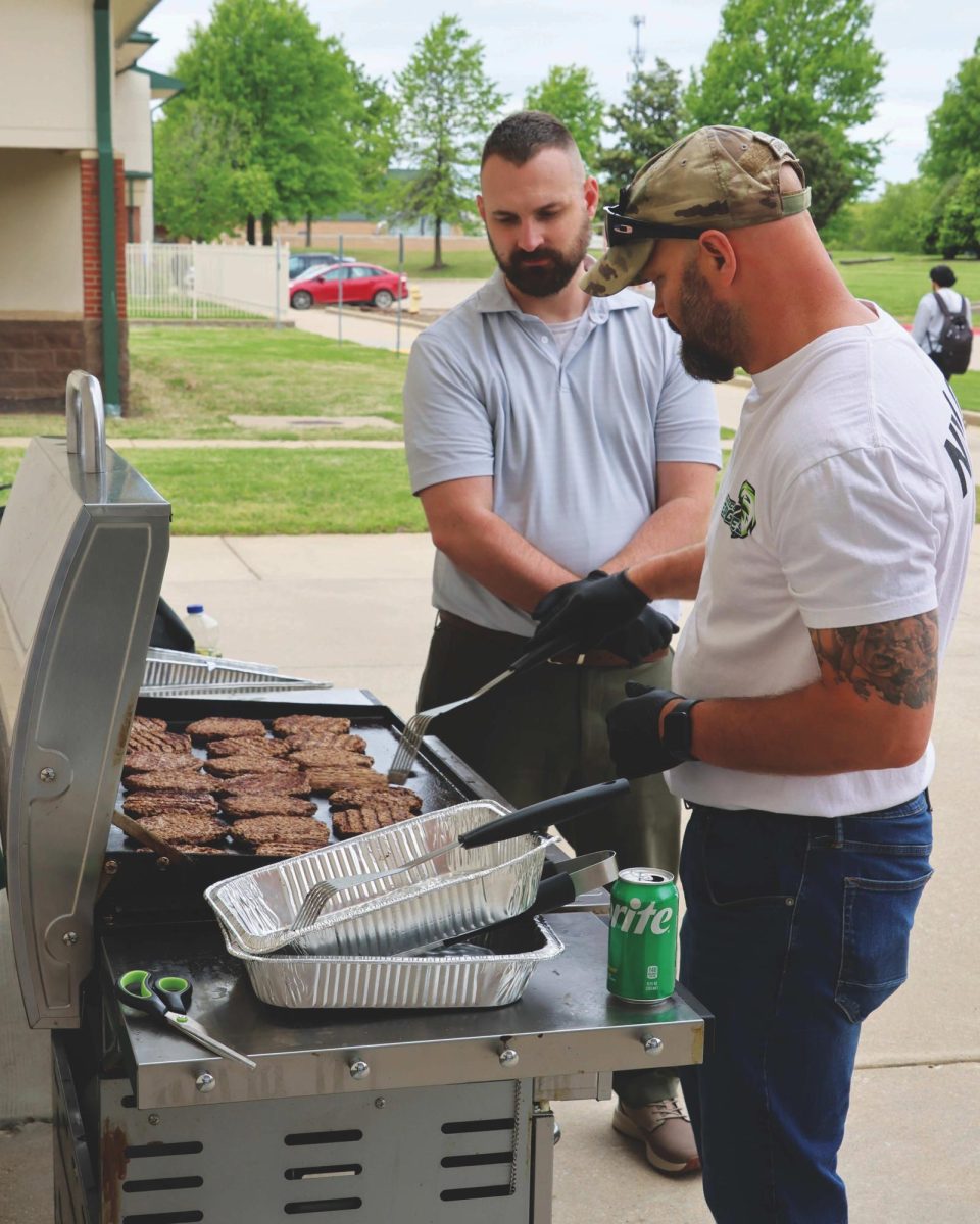 Two men stand before a barbecue grill that has hamburger patties cooking. The person on the right is using a spatula to position or test a burger.