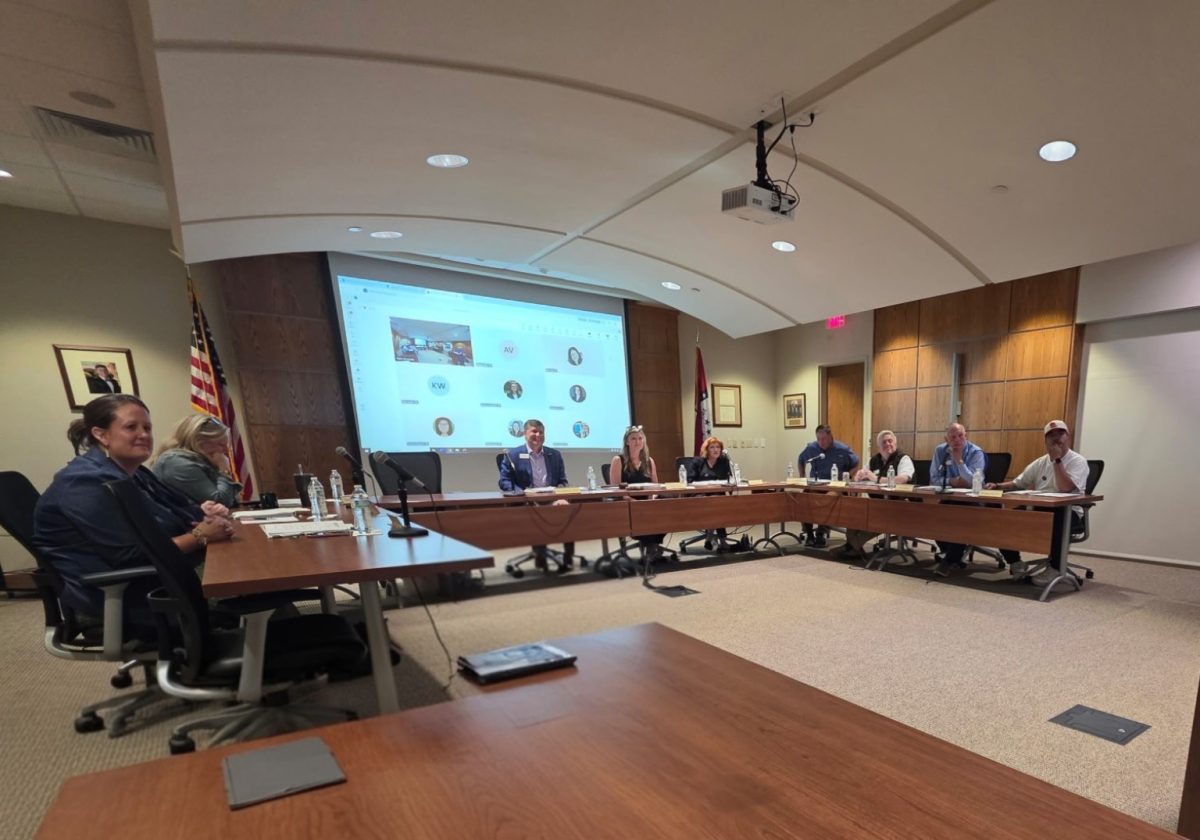 People are seated around tables arranged in a U shape. There is a screen behind them that shows images of people joining by Teams.