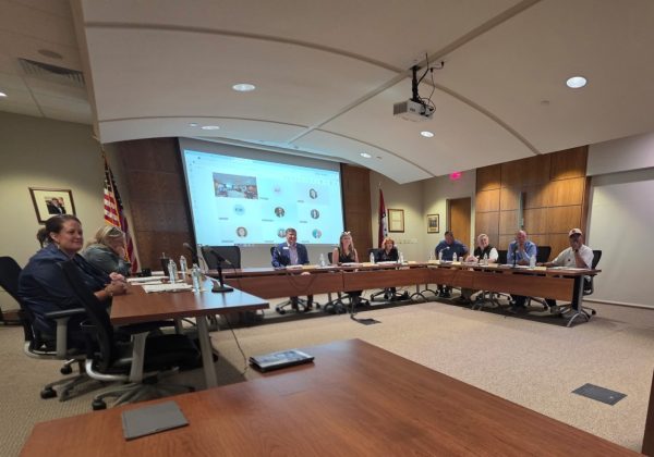 People are seated around tables arranged in a U shape. There is a screen behind them that shows images of people joining by Teams.