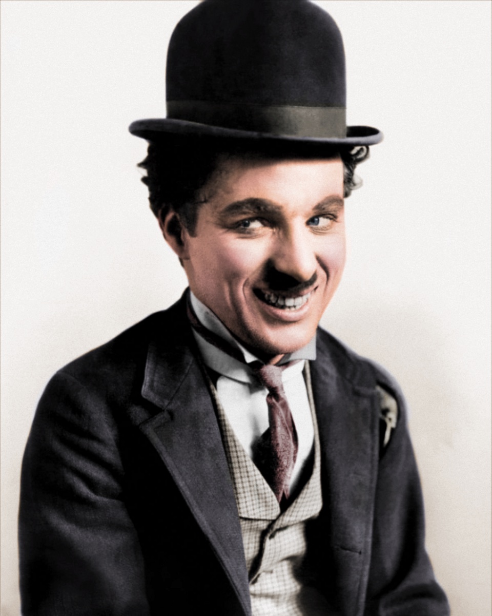 Image of Charlie Chaplin wearing a hat, shirt, tie, vest and overcoat. He is smiling and looking toward the side.