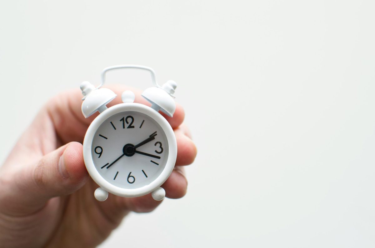 A person's left hand holds a small analog clock with the hands indicating approximately 2:17. The hands are visible and the clock face only shows numbers for 12, 3, 6 and 9.