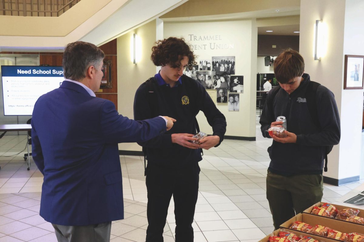 A man on the left is pointing toward a package being held by a student. A second student is look at the packaging on a pastry.