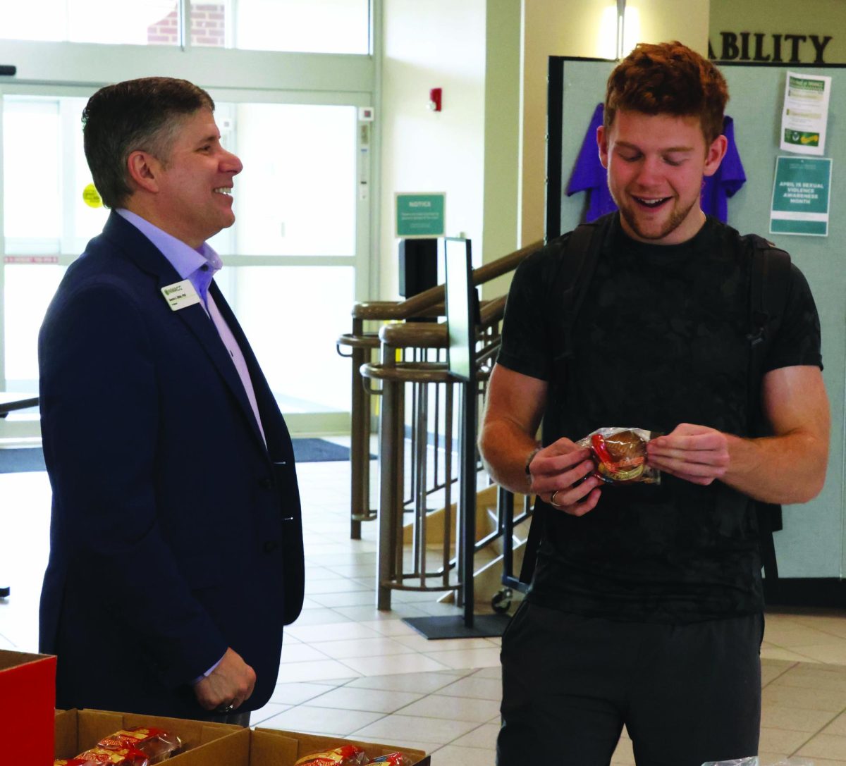 A man stands with a student who is holding a pastry package. Both are laughing or smiling.
