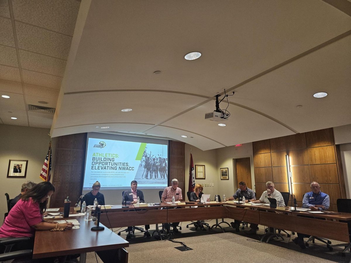 A group of people sits around a U-shaped configuration of tables. On the screen behind the center table is a projection of a slide reading, "Athletics: Building Opportunities, Elevating NWACC."